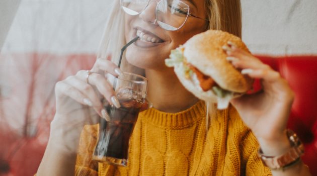 Smiling woman holding a burger, a drink, with fries on the table